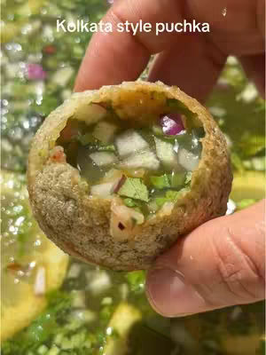 Close-up of a partially eaten puchka (pani puri) held by a hand. The puchka is filled with a mixture of chopped vegetables and a clear, watery liquid. The background is a blurred array of greenery and other puchkas.