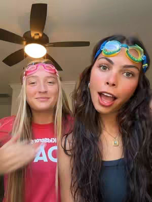 Two young women are smiling and looking at the camera, wearing swimming goggles on their foreheads. The woman on the left is wearing a red t-shirt and the woman on the right has long dark hair. The background includes a ceiling fan. The composition is well-lit with a bright, casual atmosphere.