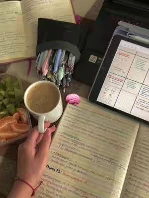 The image shows a desk setup for studying. A hand holds a cup of coffee, with a book and notes visible. There's also an open notebook with handwritten notes and some fruit. A tablet with a calendar is in the background with more books and pens.