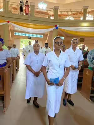 The image captures a group of women, likely nuns or religious figures, inside a bright and spacious church. They are walking towards the viewer in the middle of the aisle, showcasing a blend of architectural details and the solemnity of the scene. The lighting is bright and natural, highlighting the white attire and the wooden pews.