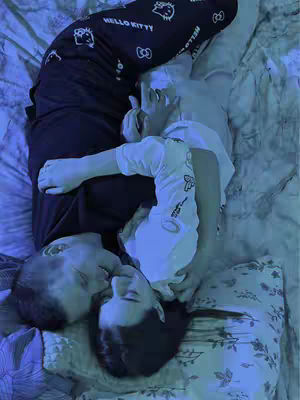 A couple is lying in bed, embracing in a tender hug. The image is bathed in cool tones, creating a serene and intimate atmosphere. One person wears a white t-shirt and the other is in black Hello Kitty pajama pants. They lie on a bed covered with a patterned bedspread and pillows.