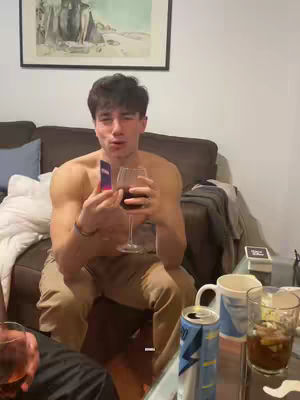 A shirtless man is sitting on a brown couch, holding a glass of red wine and a small card, with drinks and other objects on a glass table in front of him.