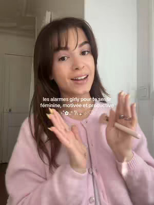 A woman with brown hair and bangs is centered in a selfie-style shot, smiling with her open hands. Behind, a room with a white door and wall is visible. The woman wears a light pink cardigan and a necklace. Text overlay is placed above the center.