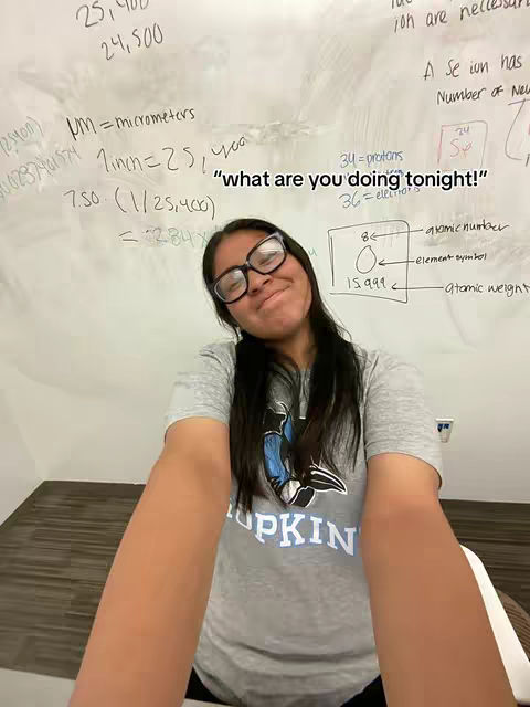 A young woman with glasses smiles at the camera, framed in a selfie-style shot. She is wearing a grey t-shirt with the word "HOPKINS" visible. The background is a whiteboard covered with equations and diagrams, indicating a classroom or study environment. The image has a casual, study-related vibe.