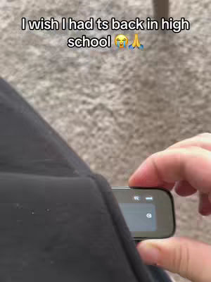 A person's hand is holding a black electronic device with a screen. The device is being held near the person's leg, which is covered by black clothing. The background consists of a textured surface, possibly carpet. The angle of the camera makes it difficult to discern the gender of the person.
