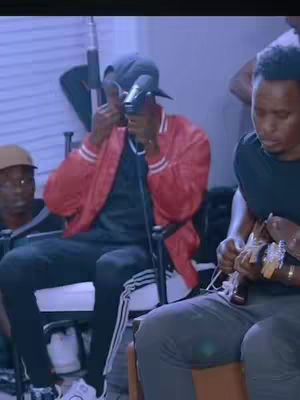 The image shows a group of men in what appears to be a recording or rehearsal setting. One man in the center, wearing a red jacket, dark cap, and baseball cap, is holding a microphone and giving a thumbs-up gesture. Around him, other men are visible, some appearing to play instruments or sing. The color palette is muted, with a cool, blueish tint. The composition is informal, capturing a moment during a performance or practice session.