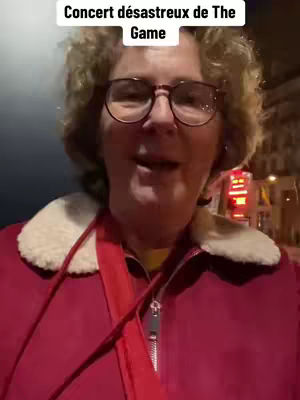 A woman wearing glasses and a red jacket stands outdoors, looking directly at the camera and speaking. The background is blurred, with hints of buildings and city lights.