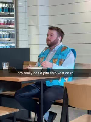 A man wearing a blue vest that seems to be made of pita chips sits at a table in a restaurant. The background includes the interior of a restaurant with a beverage fridge and an empty chair in the background.