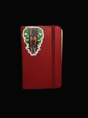 A red sketchbook or journal is centered against a stark black background. A sticker is placed on the front cover, featuring an artistic illustration of what looks like a person sitting in a chair with a gray pillar behind them, the sticker has a green border with a white outline. The cover is smooth, with a simple elastic closure along the right edge.