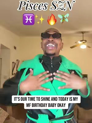 A medium shot shows a Black man in a green and black outfit with sunglasses, arms open, and mouth open in a smile. The background is a blurred living space.