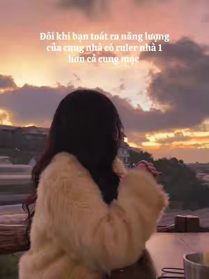 A person, seen from behind, is wearing a cream-colored fur coat. The background is a blurred sunset with shades of pink, orange, and yellow, with clouds and hints of buildings and trees. A dark countertop or surface is in the foreground, and the person's hair is dark.
