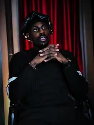 A man wearing a black hat and black outfit is sitting in a chair, with his hands clasped. Behind him is a red curtain.