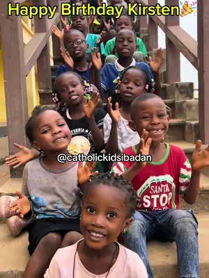 A group of smiling children, mostly of African descent, are gathered on a staircase. The children are waving at the camera. The staircase is wooden, and the overall scene feels joyful and celebratory. The text overlay says "Happy Birthday Kirsten".