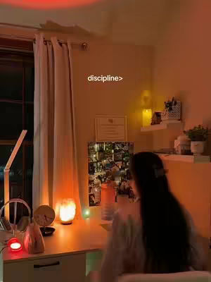A dimly lit study space with a desk, lamp, and a person working at the desk. The main colors are warm tones, the desk has a lamp and other accessories and the person is blurred out and facing away from the camera.