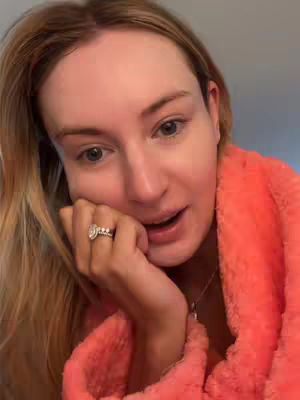 A close-up shot of a woman in an orange robe looking at the camera and smiling with her hand on her face. She has blond hair and is wearing a large diamond ring. The background is a wall.
