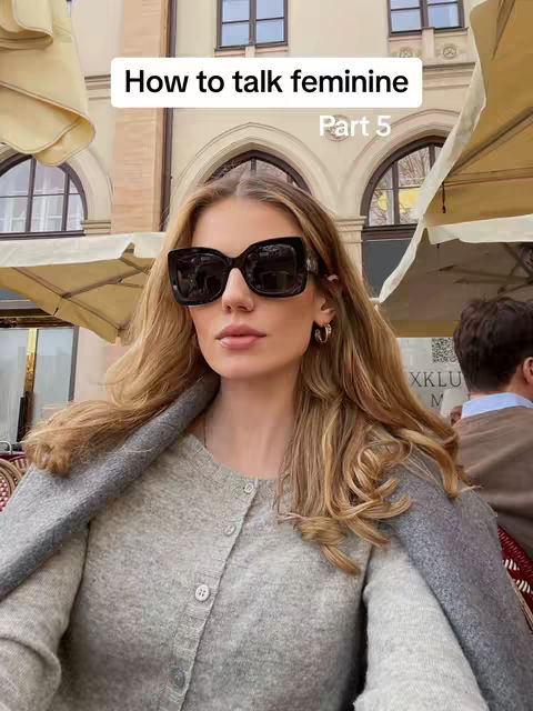 A woman with long blonde hair is wearing large black sunglasses and a gray sweater. She is sitting outdoors with buildings in the background, some of which are visible behind her. The image is brightly lit, and the woman appears to be the central focus.