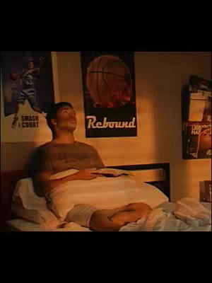 A young man lies in bed, propped up on pillows. He is looking upward with a contemplative expression. The room is softly lit with posters of a basketball and a baseball player on the wall. The lighting and setting suggest a personal space or bedroom, creating a relatable and intimate atmosphere.