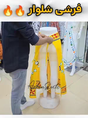 A man in a black shirt and gray jeans is holding up a yellow Farshi salwar over a dress form. The salwar has decorative patterns The background of the image shows a clothing store. Text in the image says "Farshi Shalwar" in Urdu.