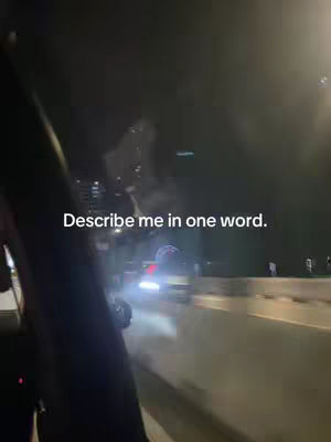 A nighttime scene from a car's interior. The camera looks out the side window, capturing the blurred motion of oncoming traffic's headlights. The text 'Describe me in one word' is superimposed on the lower portion. The overall tone is dark and enigmatic because the visibility is significantly reduced due to the darkness.