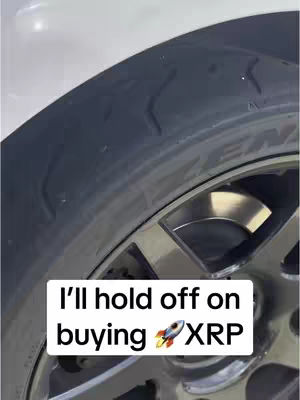 The cover image shows a close-up view of a car tire and a portion of the wheel. The black tire is angled towards the upper-left, while the wheel, with its silver finish, takes up the lower right. Overlaying this is a white rectangular text box containing the text: 'I'll hold off on buying 🚀 XRP.' The rocket emoji is positioned near the letters XRP.