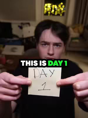 A man holds up a sticky note that says 'DAY 1' on it in front of him. The background is blurred, but it appears to be a room with some lights.