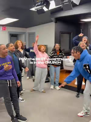 The image shows a group of people, likely in a studio setting, with various individuals singing and gesturing enthusiastically as if in a choir or vocal rehearsal. The scene is lit by overhead studio lights, and there's visible text overlayed on the image.