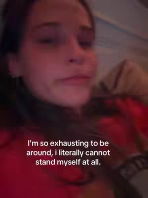 A selfie-style shot of a young woman indoors. The woman is positioned slightly off-center with her face taking up most of the frame. She has brown hair and is looking downwards with a somber expression. A red garment is visible around her shoulders and chest. Text overlayed in white at bottom.