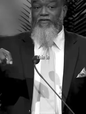 A black and white shot of a man speaking into a microphone. The man is wearing a suit, a white shirt and a tie. The man has a full, white beard.