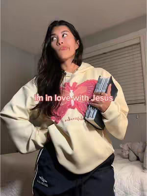 A young woman is posing in a bedroom, looking upwards with a thoughtful expression. She wears a cream hoodie with a large pink butterfly design and black athletic shorts. She holds a book, likely a Bible, and the overlaid text reads, 'im in love with Jesus'. The background shows a bed and a window with blinds.