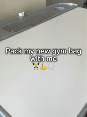 The image shows a white surface with a text overlay. The text reads, "Pack my new gym bag with me" and includes emojis of a weightlifter, a flexing bicep, and headphones. In the background, there is a hint of a shelf.