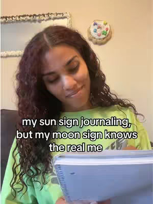 A woman with long, curly auburn hair is seated, holding a notebook and pen, smiling slightly. The background is a neutral-toned wall with a decorative mirror and a small wall decoration. Text overlayed on the image reads, "my sun sign journaling, but my moon sign knows the real me."