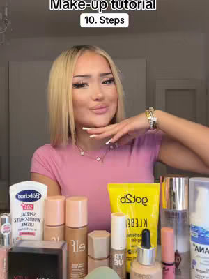 A woman with long blonde hair is in a room with makeup products and holds her hand up. The woman is wearing a pink shirt and jewelry. Text overlayed at the top indicate "Make-up tutorial" and "10. Steps".