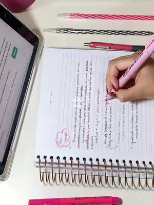 A top-down view shows a workspace with a tablet, notebook, and assorted pink pens. The notebook is open, displaying handwritten notes with diagrams and text highlighted in pink. The overall composition is bright, with white surfaces and a clean aesthetic.