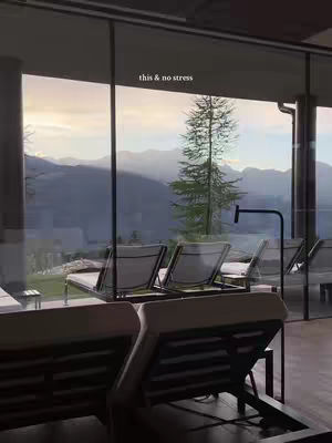 The image shows an indoor area with large windows offering a scenic view of mountains and a partly cloudy sky at dusk or dawn. Inside, there are several lounge chairs, suggesting a spa or relaxation setting. The color palette is soft, with natural light and muted tones. The composition highlights the contrast between the indoor space and the expansive outdoor vista.
