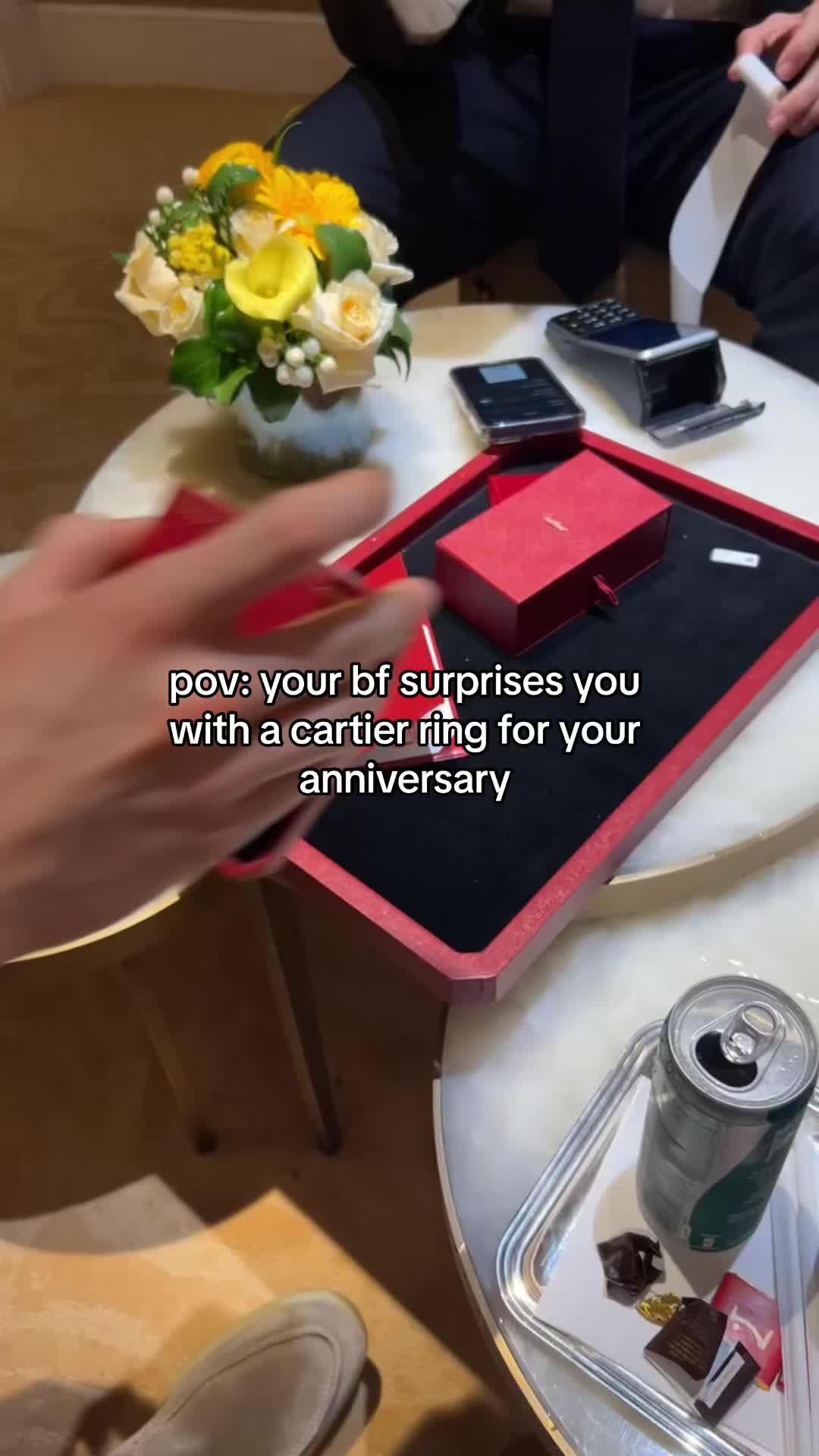 The image shows a person's hand holding a red Cartier box over a tray with a red interior. On a table with a white surface, there is a ring box, a black payment terminal, and other items like a small bouquet of flowers.