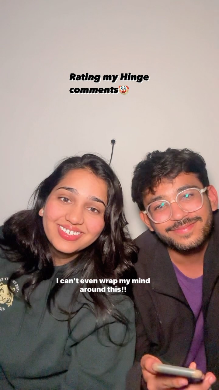 A selfie-style image featuring a young woman and a young man, both smiling and looking at the camera. The woman is on the left, with long, dark hair and a light-colored complexion. She is wearing a dark green hoodie. The man is on the right, wearing metal-rimmed glasses and a dark jacket over a purple shirt. Behind them is a plain white wall. Text overlays are present at the top and bottom of the image, with a playful emoji.