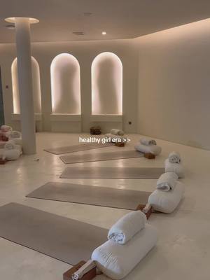 A brightly lit, minimalist room with a yoga studio setup. Neutral tones dominate the space, with rows of yoga mats, pillows, and rolled towels neatly arranged. The background features arched alcoves, and the overall aesthetic is clean and serene.