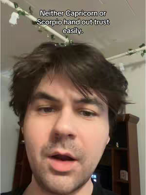 A man with short brown hair and a slight beard is centered in a selfie-style shot. The background is blurred with a glimpse of a wooden cabinet and a white ceiling trim decorated with faux vines. Text overlay on the image reads: 'Neither Capricorn or Scorpio hand out trust easily.'