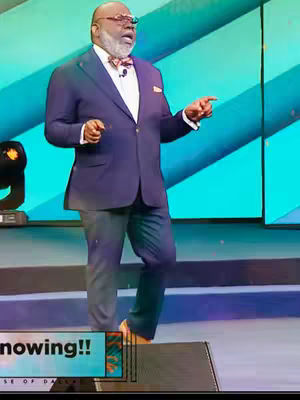 The image shows a man in a navy blue suit, white shirt, and a bow tie, addressing the audience in a speaking engagement. He is standing on a stage with a microphone, gesturing with his right hand towards the right.