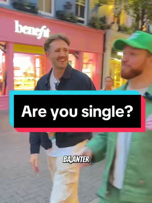 Two men are walking down a street, one wearing a navy jacket smiling while the other is wearing a green hat and jacket. Text overlayed on the image reads 'Are you single?' with the logo of the channel at the bottom.