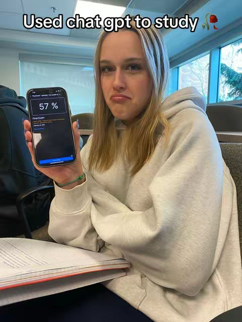 A young woman with blonde hair in a white hoodie holds up a smartphone displaying a 57% grade on a final exam. She has a somewhat distressed expression. In the background are other people and a window showing trees and sky.