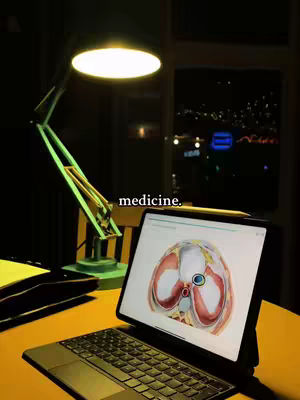 A dimly lit image showing a desk with a laptop and a table lamp. The laptop displays a medical diagram of a human torso, and the text 'medicine.' is overlaid on the image, suggesting the content is related to medical studies.