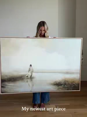 A woman stands holding a large rectangular painting. The painting depicts a figure walking toward the right in a misty, water-like landscape. The woman is blonde, wearing a white shirt and jean shorts, looking down with an admiring expression at the painting. The setting is a room with a plain white wall and wooden floors.