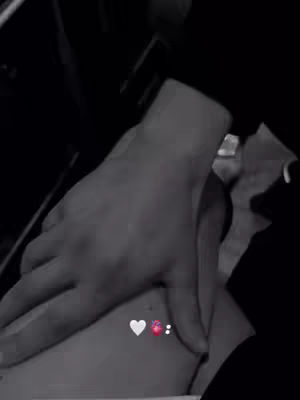 A black and white image showing two hands in an embrace. The upper hand, likely belonging to a man, is placed on the lower hand, which appears to be resting on a person's body. A heart-shaped emoji is present in the bottom center.