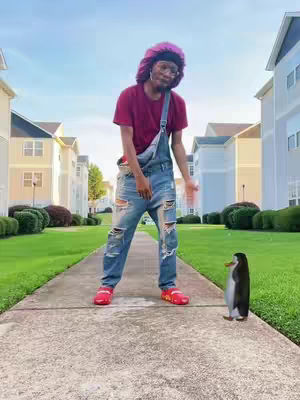 A person wearing a pink bonnet and blue jeans stands on a concrete walkway, looking towards a penguin on the right. The person is outside, with buildings and sky providing the background. The person is in a relaxed pose, possibly dancing. The penguin faces the person.