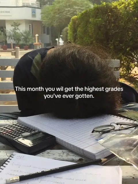 A person is leaning over a table covered in school items, with the back of their head, covered in dark hair, is the central focus. On the table are notebooks with handwritten notes, a calculator, and a pair of glasses. A pencil lies on the notebook. The background shows an outdoor setting, possibly a school campus with a building and trees.