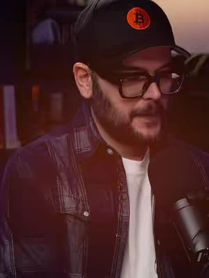 A man with a beard and glasses wearing a black cap with a Bitcoin logo, plaid shirt, and white t-shirt, sitting in front of a dark background with a microphone.