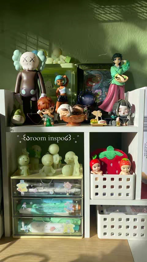 A well-lit photo displays a collection of figurines and small items arranged on a white shelving unit against a light green wall. The shelves are filled with a variety of figurines, including cartoon characters and artistic sculptures, along with decorative items like a miniature bowl and small containers. Below, there are drawers containing pencils and pens along with some miniature figurines. The lighting is natural, and the arrangement suggests an aesthetic and organized display.