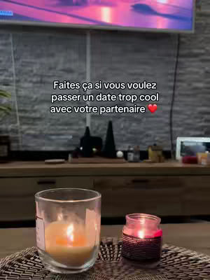 The cover image shows a cozy indoor scene, likely a living room. In the foreground, a candle in a clear glass and a smaller candle in a red jar are placed on a woven mat. Behind them, a dark wooden console table supports decorative items and a screen displays a blurry image. On the wall is some text in white font asking the audience's opinion with a red love sign at the end.