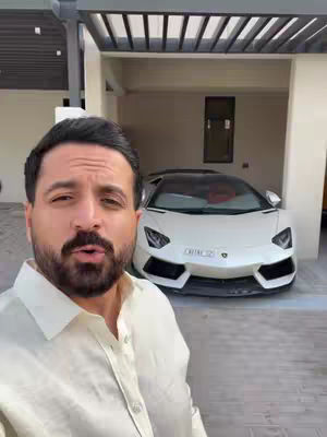A man stands in front of a white Lamborghini parked in a garage. The man, who appears to be Arab due to his facial features and hairstyle, is wearing a cream-colored button-down shirt. The car is the focal point, positioned centrally within the garage. The garage itself has a modern design, indicated by the architectural details and the dark wood slat roof that extends over the garage door. The scene is well-lit, possibly from natural light entering the garage space.